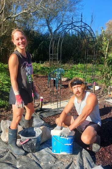 A smiling woman and a man work together outdoors in a garden, preparing materials with gloves and boots, under a bright blue sky and metal arch.