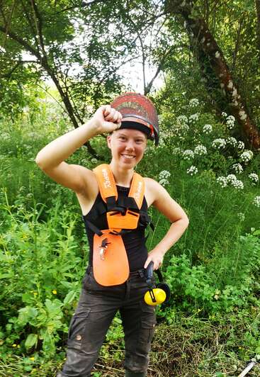 A smiling person in protective gear, including a visor and orange harness, stands outdoors in lush greenery, holding ear protection, appearing ready for outdoor work.
