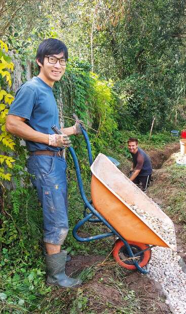 Dos hombres trabajan al aire libre en una zona verde y frondosa. Uno de ellos, que lleva guantes, sostiene una carretilla llena de piedras, mientras que el otro cava y sonríe.