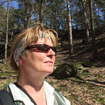 A imagem mostra uma mulher em pé em uma área arborizada, usando óculos escuros e uma faixa na cabeça, com árvores e rochas visíveis ao fundo em um dia ensolarado.