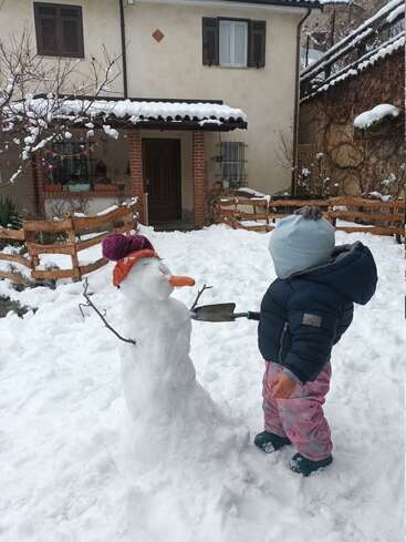 Un niño pequeño vestido con ropa de invierno está de pie junto a un muñeco de nieve con gorro y bufanda, y nariz de zanahoria, en el jardín nevado de una casa.