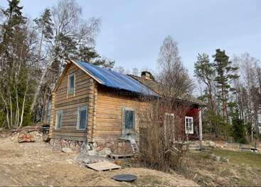 Une maison rustique en bois, aux fondations en pierre, se dresse dans une clairière à la campagne, entourée d'arbres dénudés et de conifères, évoquant le début du printemps ou la fin de l'automne.