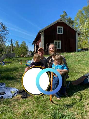 Une joyeuse famille de quatre personnes est assise sur l'herbe verte devant une maison rustique en bois rouge ; ils profitent ensemble d'une journée ensoleillée, tenant dans leurs mains des objets ronds et colorés, et arborant un sourire chaleureux.
