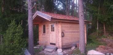 Une petite cabane en bois au toit de tuiles rouges se dresse au milieu de grands pins. Nichée au cœur de la forêt, elle se distingue par sa porte et sa fenêtre sobres, ainsi que par son charme rustique.