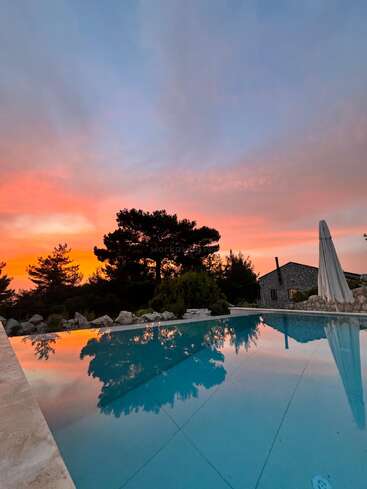 Un magnifique coucher de soleil aux teintes orange et bleues vibrantes se reflète magnifiquement dans une piscine tranquille, entourée d'arbres, de rochers et d'une maison en pierre.