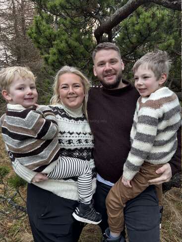 Une joyeuse famille de quatre personnes se tient à l'extérieur, les parents tenant chacun un enfant dans leurs bras. Ils portent des pulls confortables à motifs, arborent un sourire chaleureux et sont entourés de pins et de nature.