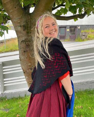 Une jeune femme souriante aux longs cheveux blonds se tient à l'extérieur, sous un arbre, vêtue d'un costume traditionnel composé d'un châle rouge et noir et d'une jupe rayée.