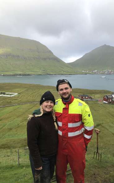 Un couple souriant se tient sur une colline verdoyante surplombant un lac et un petit village, entouré de montagnes sous un ciel nuageux. L'homme porte un costume brillant et réfléchissant.
