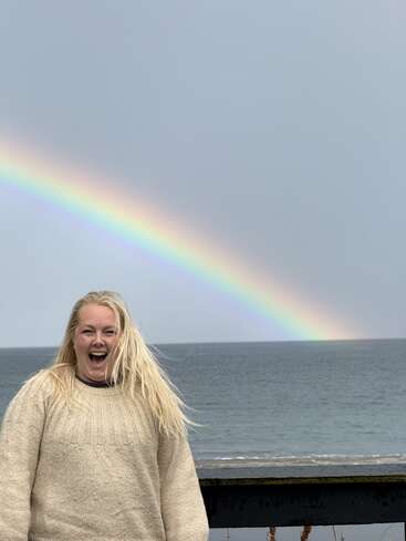 Une femme aux cheveux blonds se tient au bord de l'océan, le sourire aux lèvres. Derrière elle, un arc-en-ciel aux couleurs vives se dessine dans un ciel nuageux, apportant de la couleur et de la joie à la scène.