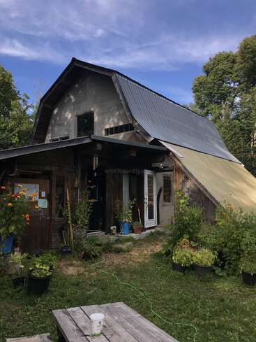 Una casa rústica con tejado metálico inclinado se alza entre la vegetación. La entrada está decorada con macetas y flores. En primer plano hay una mesa de picnic.