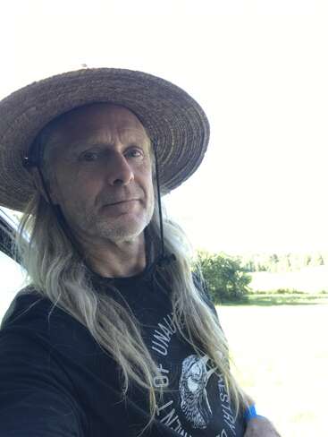 Un hombre de pelo largo y rubio lleva un sombrero de paja de ala ancha y una camiseta gráfica negra. Está de pie al aire libre en un día soleado con vegetación detrás.