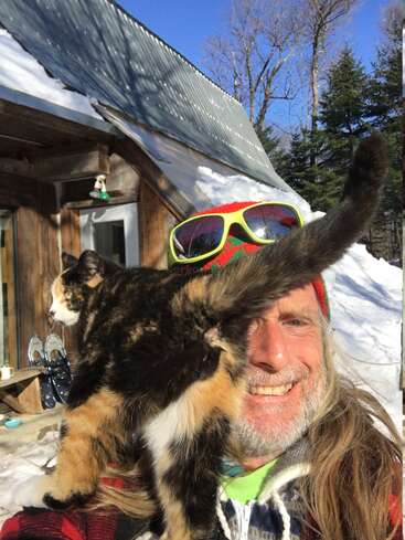 La imagen muestra a un hombre de pelo largo y sombrero de colores, sonriendo a la cámara, con un gato posado en su hombro frente a un edificio nevado.