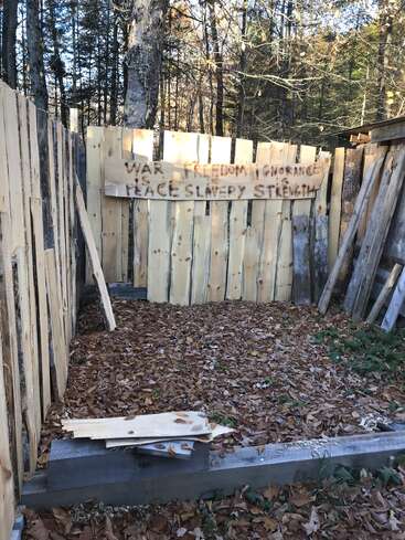 Uma cerca de madeira improvisada em uma clareira na floresta, com folhas de outono no chão. Uma placa diz: "GUERRA É PAZ LIBERDADE É ESCRAVIDÃO IGNORÂNCIA É FORÇA".