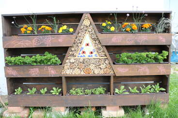This image shows a wooden garden planter with multiple sections, colorful flowers, herbs, and a central triangular insect hotel, adorned with insect drawings and mosaic details.