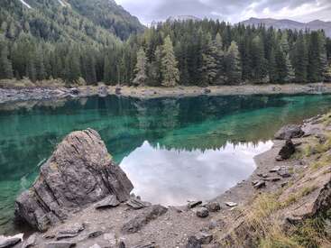 Un lac turquoise, serein et cristallin, entouré d'une forêt dense à feuilles persistantes, de rivages rocheux et de montagnes lointaines sous un ciel nuageux, reflétant la beauté naturelle et la paix.