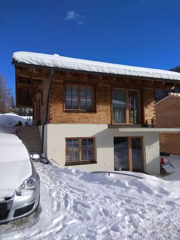 Une maison en bois dont le toit et la cour sont recouverts de neige se dresse sous un ciel bleu clair. Une voiture et une luge argentées sont partiellement enfouies dans la neige fraîche à l'extérieur.