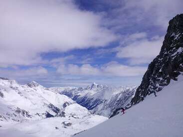 Un magnifique paysage d'hiver avec des montagnes enneigées sous un ciel partiellement nuageux. Deux skieurs descendent une pente raide et enneigée à côté d'un affleurement rocheux.
