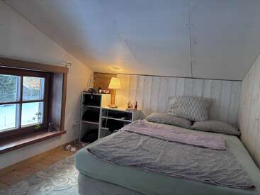 Cette chambre mansardée confortable est dotée d'un plafond incliné, d'un lit avec des oreillers et des couvertures, d'une petite table de nuit avec une lampe, d'une étagère et d'une fenêtre.