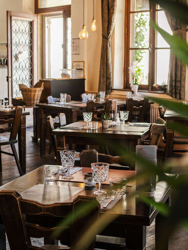 Cette image montre un restaurant confortable et ensoleillé, avec des tables et des chaises en bois, magnifiquement dressées avec des verres en cristal, des serviettes, des couverts et un éclairage décoratif chaleureux. Une atmosphère accueillante.