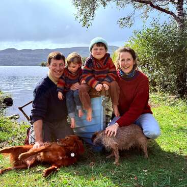 A happy family of four, wearing cozy sweaters, poses by a lake with two dogs. The scenic background includes water, trees, and distant hills.