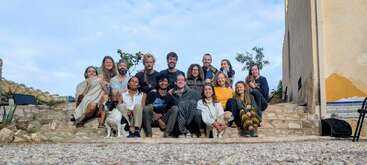 Eine Gruppe von sechzehn Menschen und zwei Hunden sitzt gemeinsam auf einer Steintreppe im Freien und lächelt in die Kamera. Die Szene ist hell und freundlich und suggeriert Zusammengehörigkeit.