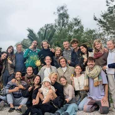 A joyful group of people pose outdoors, smiling and holding yellow flowers. There are lush green trees and palms behind them, creating a warm, friendly atmosphere.