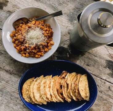 Sobre una mesa rústica de madera hay un cuenco con patatas cortadas en dados y queso, un plato con tortillas dobladas y un termo de metal, lo que sugiere una suculenta comida casera.