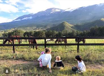 Una mujer y dos niños están sentados en la hierba, contemplando a los caballos que pastan en un pintoresco valle montañoso. El paisaje es frondoso y tranquilo, con onduladas colinas verdes.