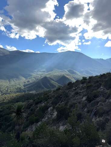This image shows a mountainous landscape covered with lush green trees, dramatic clouds in the sky, and beams of sunlight shining through, creating a peaceful scenic view.