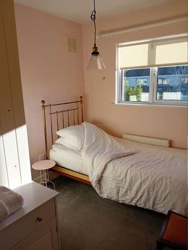 Um quarto iluminado pelo sol com paredes rosa pastel, uma cama de latão, roupa de cama branca, uma pequena mesa lateral redonda, uma janela e uma moderna luminária suspensa. Ambiente aconchegante.
