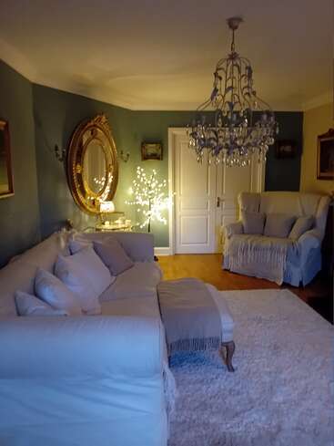 Esta sala de estar aconchegante tem um lustre elegante, um espelho grande, móveis brancos macios, tapete fofo, piso de madeira, luzes decorativas brilhantes e paredes clássicas pintadas de azul esverdeado.