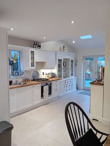 Uma cozinha moderna e bem iluminada com armários brancos, bancadas de madeira, área de jantar, vaso decorativo, garrafas de vinho, estátua de Buda e portas de vidro que levam a um pátio aconchegante.