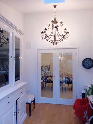 Um cômodo aconchegante e elegante com um grande lustre, portas duplas de vidro, armários brancos, piso de madeira, uma cadeira floral e um vislumbre de uma área de jantar mais adiante.