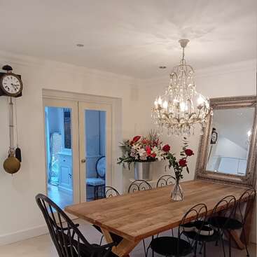 A cozy dining room with a wooden table, black chairs, a crystal chandelier, large mirror, wall clock, floral arrangements, and double doors leading to another room.