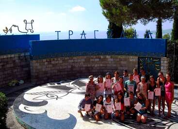 A group of young people poses in an outdoor amphitheater, holding certificates. The backdrop features Greek-inspired designs, blue walls, and the word "TRAGΩΔΙΑ."
