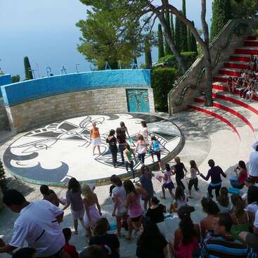 This image shows an outdoor amphitheater with people, mostly children, gathered and participating in what appears to be a group activity or dance near the stage.