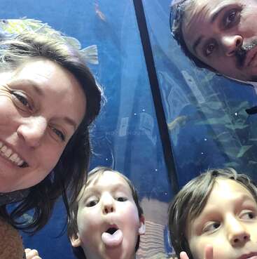 A family of four takes a fun selfie at an aquarium. The kids make funny faces; the parents smile and look happy. Fish swim behind them.