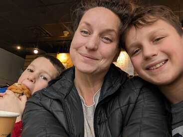 A woman smiles with two boys inside a cozy café. One boy eats a muffin, the other smiles at the camera. Warm, joyful, and casual atmosphere.