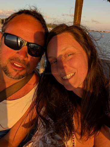 A smiling couple enjoys golden hour together by the water. The man wears sunglasses and a tank top, while the woman has loose hair and a patterned top.