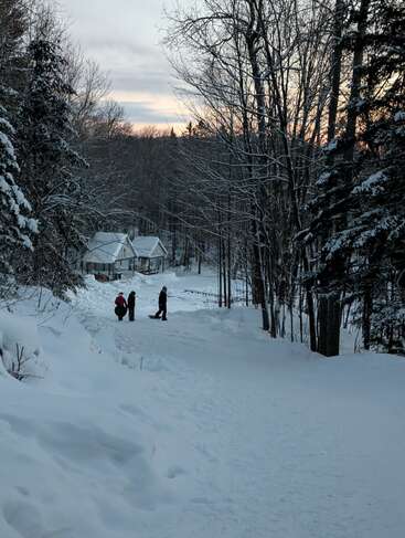 Snow covers the ground and trees as three people, bundled up, stand near a cabin in a peaceful winter forest at dusk, sky tinged orange.