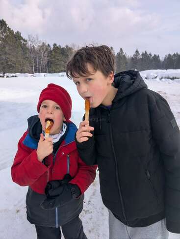 Two children, dressed in winter jackets, stand in snowy outdoors, happily licking maple taffy on sticks. Trees and snowbanks form the backdrop under a cloudy sky.