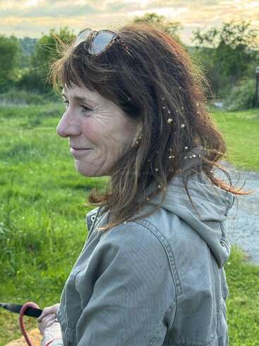 Uma mulher está ao ar livre, num campo verde, ao pôr do sol, sorrindo gentilmente. Ela tem óculos escuros na cabeça e pequenas flores espalhadas pelo cabelo.