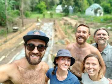 Five smiling people pose for a selfie outdoors in a garden or farm. Three are shirtless, two wear hats. Lush greenery and planting beds appear in the background.