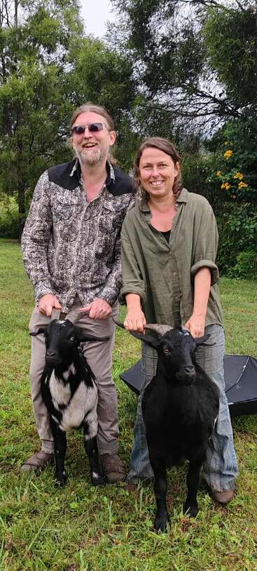 A smiling man and woman stand outdoors, each holding a goat by its horns. They're surrounded by lush greenery, giving a cheerful, rustic atmosphere on a cloudy day.
