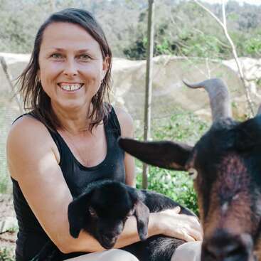 A smiling woman sits outdoors, holding a black baby goat in her arms. Another adult goat is in the foreground, with green foliage in the background.