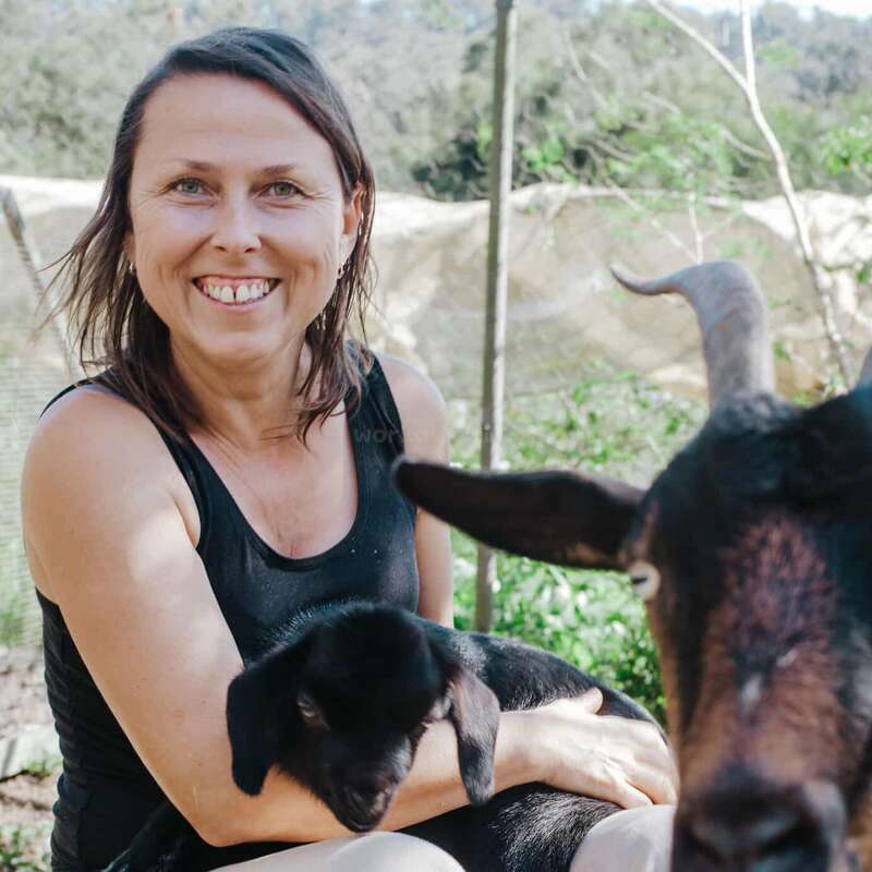 A smiling woman sits outdoors, holding a black baby goat in her arms. Another adult goat is in the foreground, with green foliage in the background.