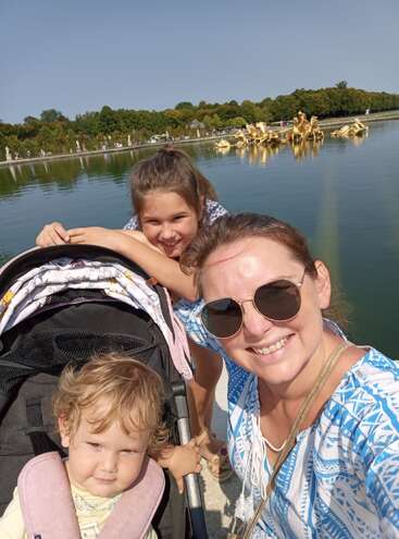 Uma mulher de óculos escuros sorri ao lado de duas crianças, uma delas no carrinho, junto a um lago espelhado com estátuas douradas e árvores ao fundo, sob um céu limpo.
