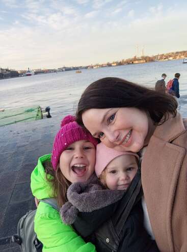 Uma mulher e duas crianças, bem agasalhadas com roupas de inverno, sorriem juntas perto da orla, com barcos e prédios da cidade ao fundo, num dia de céu limpo.