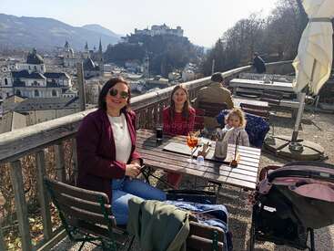 Uma mulher e duas meninas tomam um drinque em um café ao ar livre com uma vista deslumbrante da cidade e do castelo, sob um céu ensolarado, rodeadas por montanhas e árvores.
