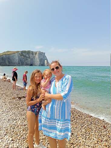 A woman and two children smile on a pebbled beach with turquoise sea, cliffs in the background, and other people enjoying a sunny summer day.